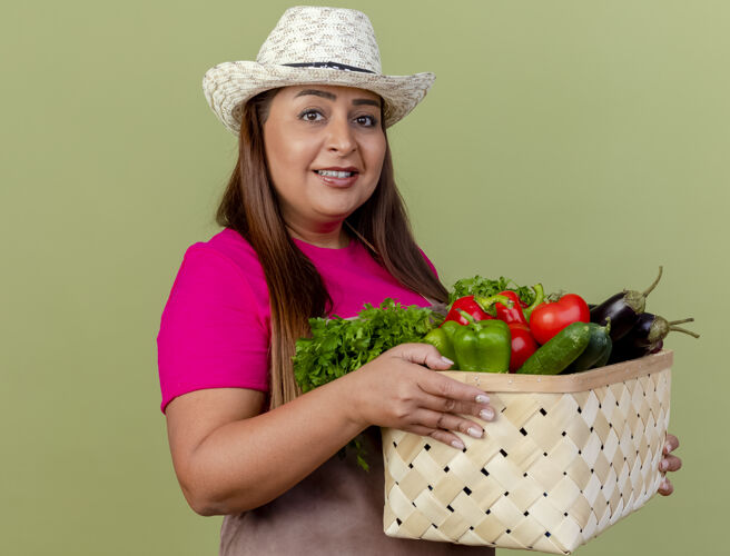 园丁中年女园丁围着围裙 戴着帽子 手里拿着装满蔬菜的箱子 站在灯光背景下 面带微笑地看着摄像机女人蔬菜抱着