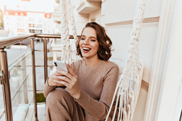 风景坐在阳台上看电话留言的好女孩阳台上兴奋可爱的女士拿着智能手机摆姿势的照片女人快乐年轻