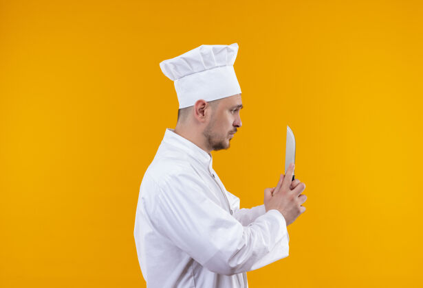 厨师年轻帅气的厨师穿着厨师制服拿着刀看着它站在偏僻的橙色空间的侧视图上帅气年轻橙色