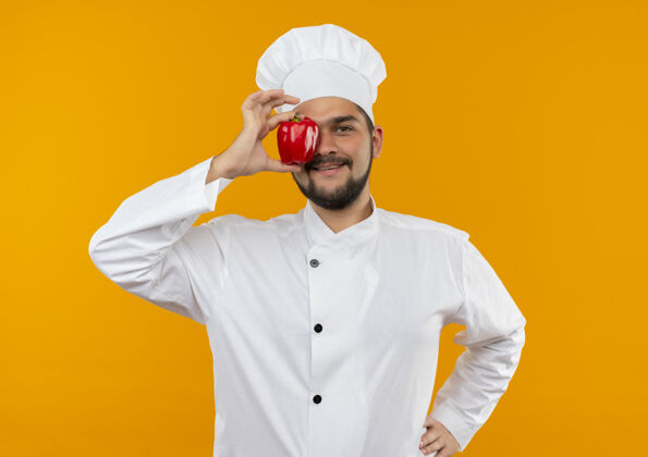 制服微笑的年轻男厨师身着厨师制服 把胡椒粉放在眼睛上 手放在腰上 孤立在橙色的空间里橘子年轻腰