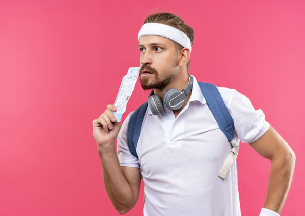 穿着体贴的年轻帅气的运动型男人戴着头带和腕带 脖子上背着一个带耳机的包 手里拿着机票 看着粉色空间里孤立的一面票复制太空
