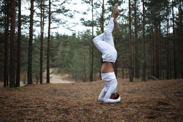 瑜伽赤脚穿着白色衣服的年轻男子在森林里的地面上做萨拉姆巴shirshasana瑜伽姿势的变化 交叉双腿在森林里高级瑜伽训练的户外镜头 双手平衡练习年轻肌肉
