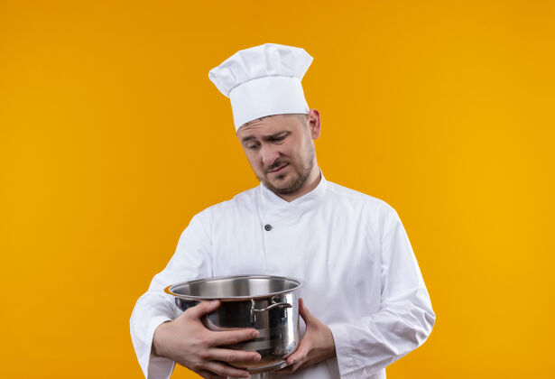 烹饪体贴的年轻帅哥厨师穿着厨师制服 拿着锅炉 看着孤立的橙色空间拿着英俊年轻