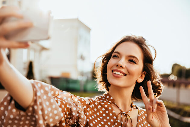 积极幸福的短发女人穿着棕色衣服表达幸福快乐的女人拿着电话自拍乐趣黑发寒冷