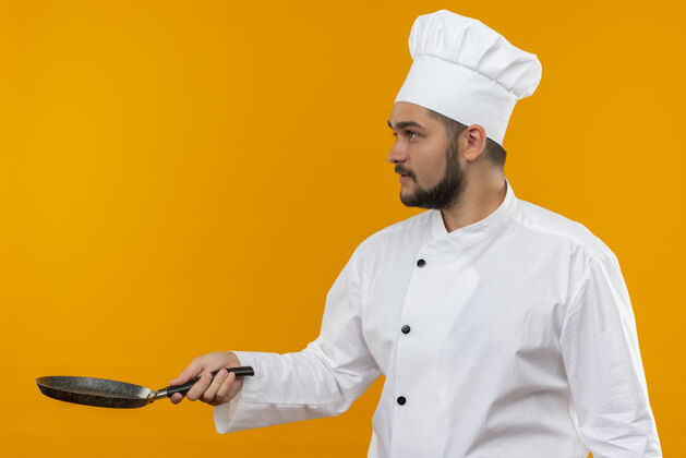 厨师身穿厨师制服的年轻男厨师手拿煎锅 看着橙色空间上孤立的一面看拿着男
