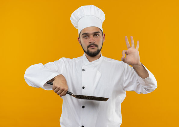 男身着厨师制服的年轻男厨师手拿煎锅 在橙色的空间里做着“ok”的手势制服做着拿着