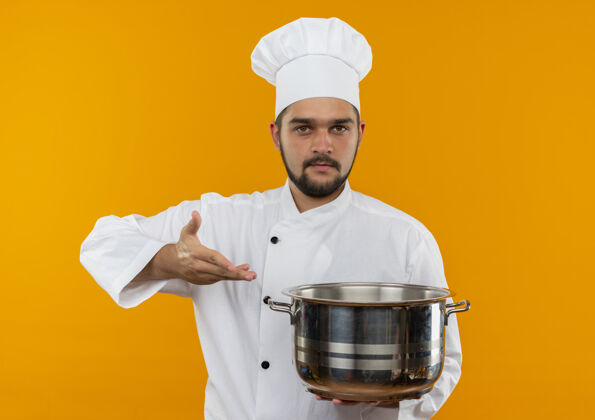 看身着厨师制服的年轻男厨师手拿手指着锅 看上去孤立在橙色的空间里厨师指着厨师