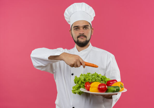烹饪身着厨师制服的年轻男厨师拿着盘子里的蔬菜 用胡萝卜指着隔离在粉红色空间里的那个盘子持有胡萝卜盘子