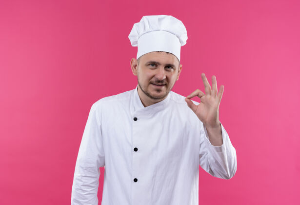 年轻高兴的年轻帅哥厨师在厨师制服做ok标志上孤立的粉红色空间招牌高兴粉红