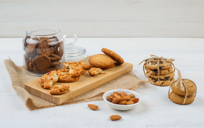 碗一些棕色的饼干和杏仁放在碗里 饼干放在砧板上 还有一个装在白色表面玻璃罐里的袋子巧克力薯条切割