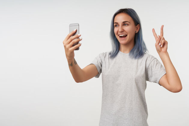 站着快乐的年轻短发女士 身上有纹身 用胜利的手势举手 微笑着在智能手机上为自己画肖像 在白色屏幕上与世隔绝表情积极欧洲