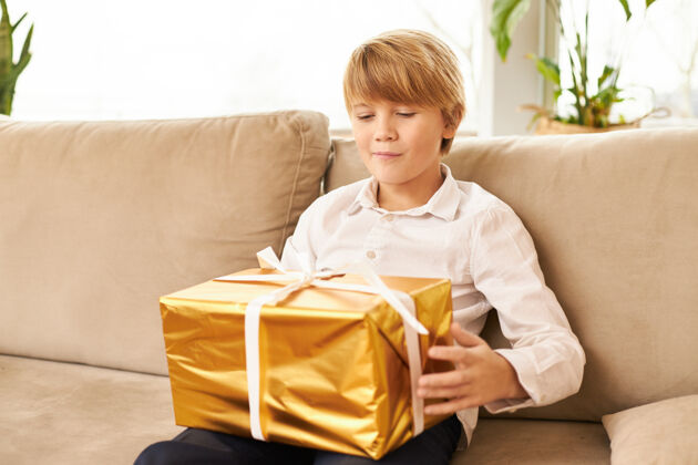 孩子可爱的白人少年坐在沙发上 膝上放着新年礼物帅哥准备打开装有圣诞礼物的金色盒子 脸上露出好奇的期待的表情 微笑着童年快乐盒子