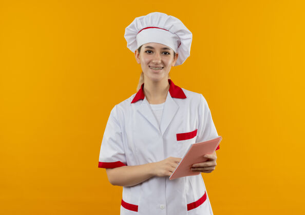 微笑微笑着的年轻漂亮的厨师 穿着厨师制服 戴着牙套 把手指放在橙色空间的笔记本上年轻漂亮牙套