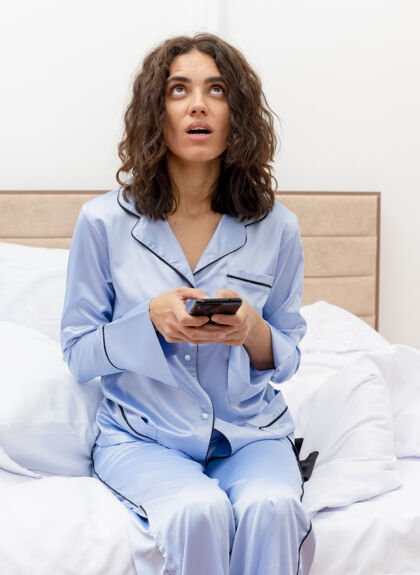 女人穿着蓝色睡衣的年轻漂亮女人坐在床上 用智能手机在卧室的室内灯光背景下困惑地仰望着困惑坐着起来