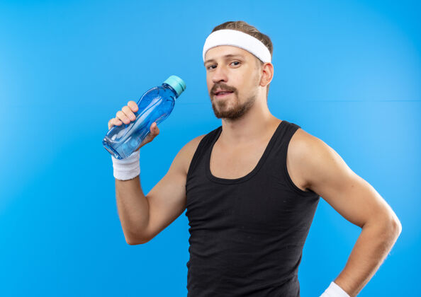 瓶子高兴的年轻英俊的运动型男子戴着头带和手镯拿着水瓶看起来孤立的蓝色空间头带蓝色帅气