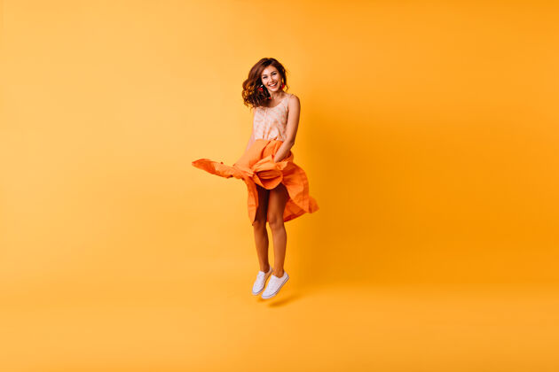 卷发温文尔雅的时尚女士在演播室跳跃的全长肖像穿着橙色裙子的漂亮姜汁女孩玩得很开心有趣现代放松