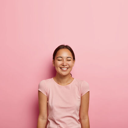 快乐情感满足的亚洲女性肖像 自然美丽 深色梳发 微笑愉快 闭上眼睛 穿着休闲t恤 隔离在粉红色的墙上人 种族 积极的情感高兴高兴穿孔
