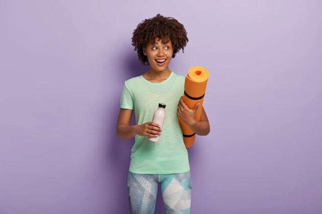 女孩开朗的黑皮肤女孩拿着健身垫和一瓶淡水 在疲惫的运动中喝着饮料 穿着运动服在室内摆出姿势积极 健康的生活方式训练女性身体