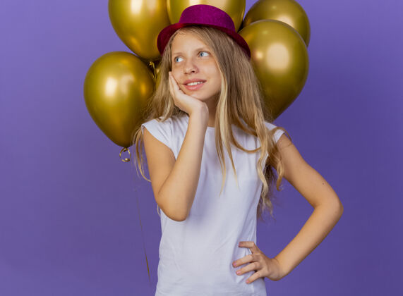 看着戴着节日礼帽的漂亮小女孩和一堆巴隆人看着一边 脸上洋溢着幸福的表情 脸上洋溢着积极的情绪 紫色背景下站着生日派对的概念紫色小感觉