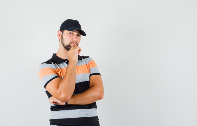 男性年轻人站在思考的姿势在t恤衫 帽子和看起来明智敏感自信衬衫