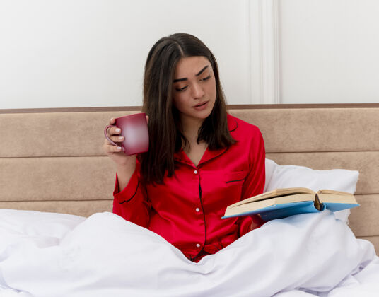 书穿着红色睡衣的年轻美女坐在床上 一边喝着咖啡 一边看书 放松地享受着周末的时光 在卧室的室内灯光背景下看书放松女人卧室