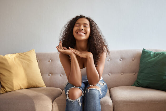 快乐一幅充满喜悦情绪的年轻黑皮肤女性的画像 她坐在客厅舒适的沙发上 穿着时髦的破牛仔裤 抬起头 双手放在下巴下 心情愉快 面带微笑沙发肖像非洲