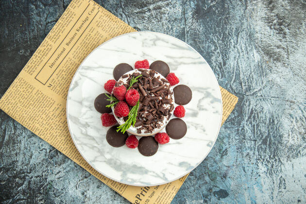 美食顶视芝士蛋糕与巧克力和覆盆子在白色椭圆形平板上报纸上的灰色表面自由空间椭圆形视图浆果