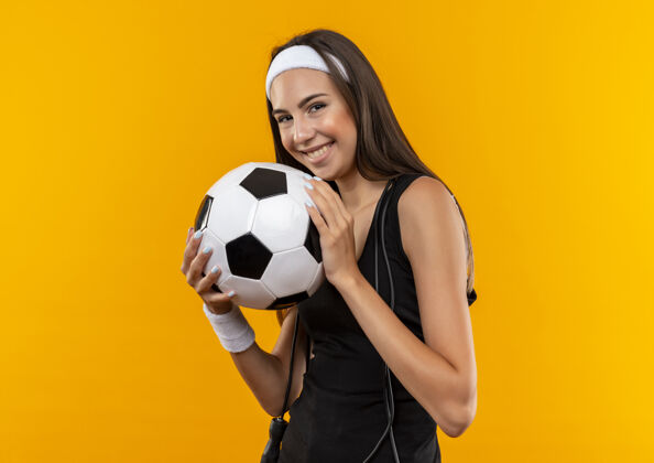足球带着微笑的年轻漂亮的运动女孩戴着头带和腕带拿着足球 脖子上系着跳绳孤立在橙色的空间里周围年轻脖子
