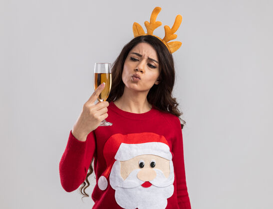 噘嘴困惑的年轻漂亮女孩戴着驯鹿的鹿角头带和圣诞老人的毛衣拿着 看着一杯香槟和噘嘴嘴唇女孩圣诞老人