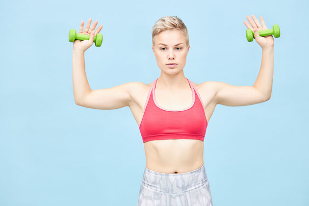健身房一张年轻漂亮的运动型女性的照片 她留着一头孩子气的头发 做着体育锻炼 双手抱着两个绿色的哑铃 侧着胳膊 训练着二头肌和肩部肌肉年轻成人体重