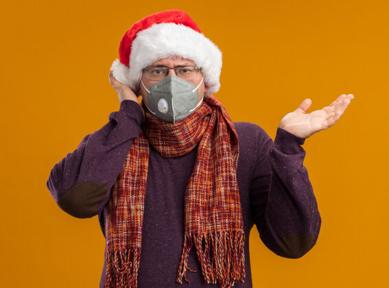 印象深刻一个戴着防护面具眼镜 戴着圣诞老人帽子 脖子上围着围巾的男人看着镜头 一只空手触摸着孤立在橙色背景上的头脖子周围帽子