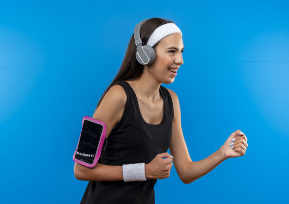 运动快乐的年轻漂亮的运动女孩戴着头带和腕带 戴着耳机和电话袖标站在侧视图看蓝色空间孤立的一面戴个人资料女孩
