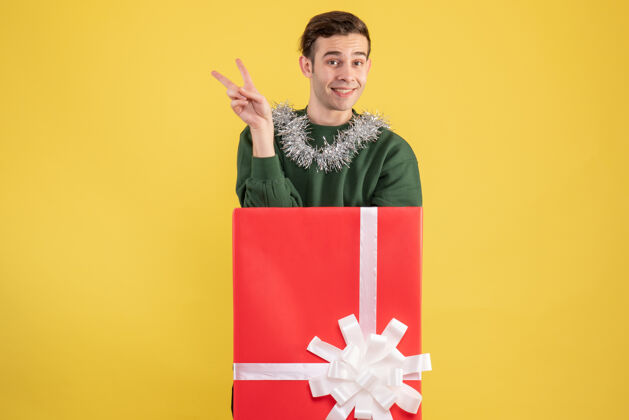 年轻人正面图：年轻人站在黄色大礼盒后面做v形标志购物礼物标志