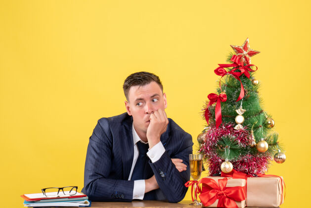 坐着前视图：体贴的男人坐在圣诞树旁的桌子旁 黄色的礼物男性公司树