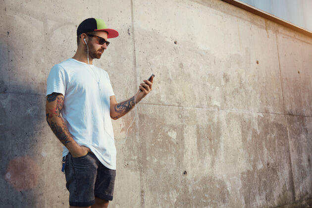 智能一个年轻人站在灰色的水泥墙旁边 看着智能手机的屏幕 戴着白色的耳塞听音乐耳机收听空白