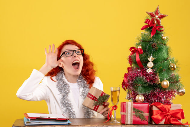 快乐前视图：女医生坐在桌子后面 黄色背景上有圣诞礼物漂亮护士坐着