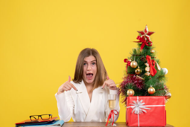 人正面图：女医生坐在黄色背景的桌子前 放着圣诞树和礼品盒漂亮成人年