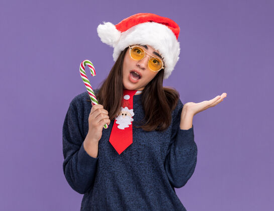 女孩焦急的年轻白人女孩戴着太阳眼镜 戴着圣诞帽 打着圣诞领带 手里拿着糖果手杖 在紫色的背景上 手一直张开 留着空白手杖举行背景