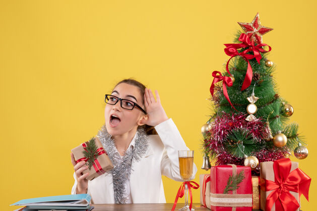 办公室前视图：女医生拿着圣诞礼物和黄色背景上的圣诞树坐着成人女性情感