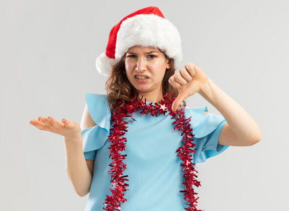 困惑身穿蓝色上衣 戴着圣诞帽 脖子上戴着金属丝的年轻女子看起来既不高兴又困惑 不高兴地竖起大拇指站蓝色手臂