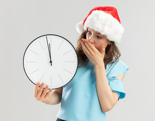 钟戴着蓝色上衣和圣诞帽的年轻女子拿着挂钟看着它被震惊用手捂住嘴巴帽子嘴年轻