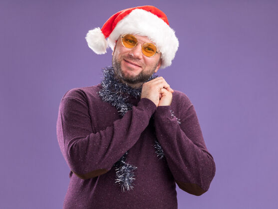 高兴戴着圣诞帽 脖子上戴着金箔花环 戴着眼镜的中年男子高兴地把两只手分开放在紫色的墙上脖子金属丝花环
