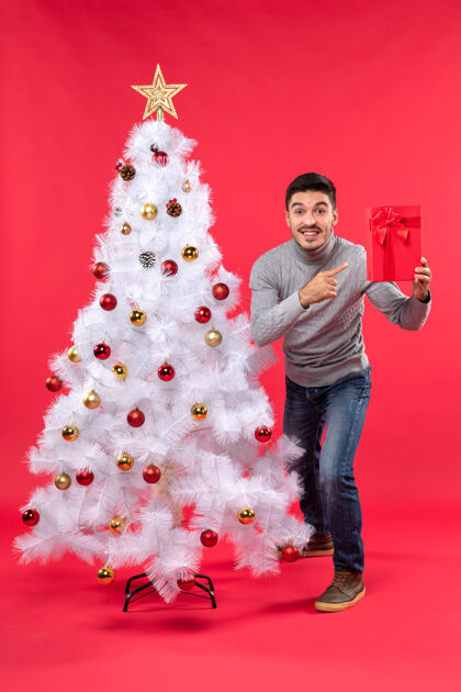 庆祝俯视图：年轻人站在装饰过的白色新年树旁 手里拿着他的礼物指着圣诞年轻人