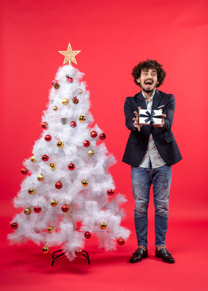 圣诞树震惊的年轻人展示他的礼物站在附近装饰白色圣诞树右侧的红色礼物一边礼物