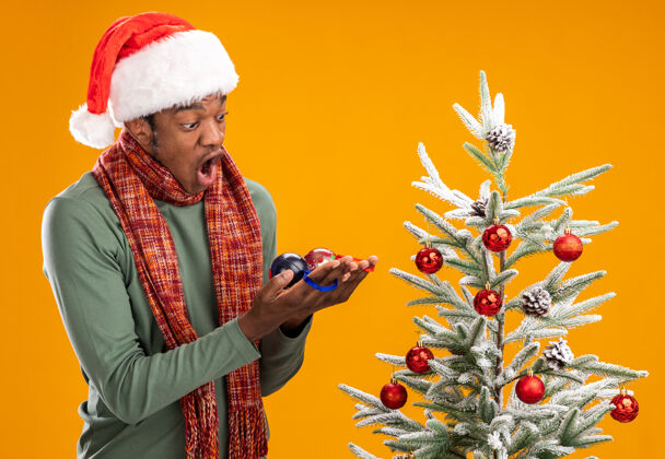 非洲人一个戴着圣诞帽 脖子上围着围巾的非洲裔美国人拿着圣诞球 站在橙色背景下的圣诞树旁惊讶地看着它们站着圣诞老人脖子