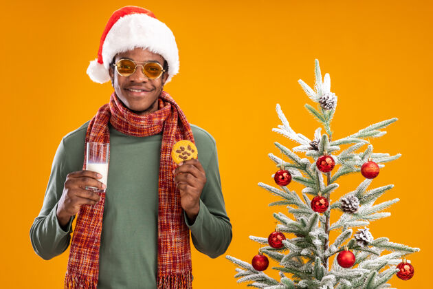 抱着快乐的非洲裔美国人戴着圣诞帽 围着围巾 手拿牛奶和饼干 微笑着站在橙色背景下的圣诞树旁脖子非洲男人
