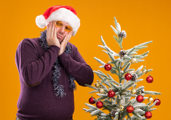 花环困惑的中年男子戴着圣诞帽 脖子上戴着金箔花环 戴着眼镜站在装饰过的圣诞树旁困惑附近穿着