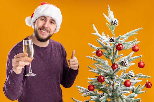 站着穿着紫色毛衣 戴着圣诞帽的年轻人在橙色背景下的圣诞树旁举着一杯香槟看展示男人