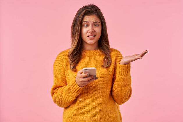 站立室内画像年轻的棕色头发困惑的女性与不高兴的脸和提高困惑的手 而站在粉红色的智能手机棕色女性表情
