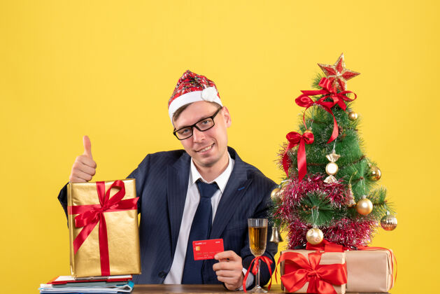 拇指商务人士在圣诞树旁的桌子旁做竖起大拇指的招牌 黄色的礼物商人成人男性